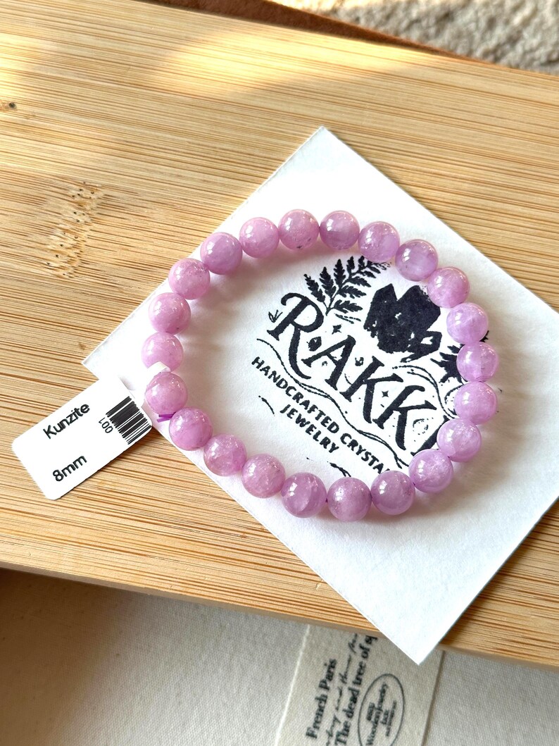 May include: A light purple kunzite bracelet with 8mm beads rests on a white paper with the brand name "Rakk" and the words "Handcrafted Crystal Jewelry". The bracelet is displayed on a wooden surface.