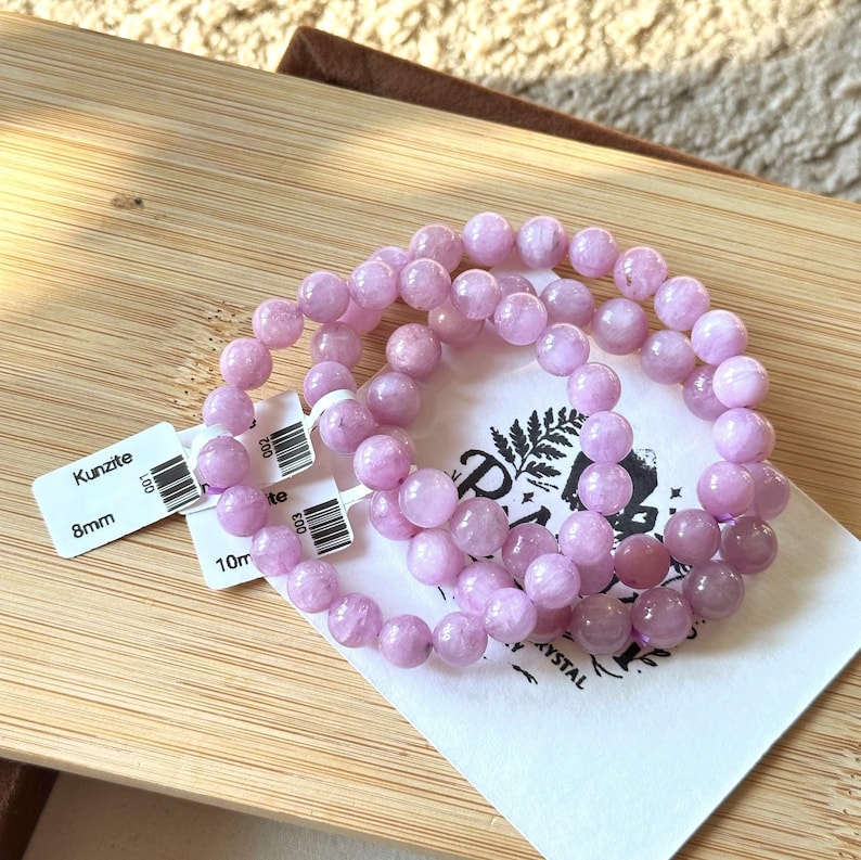 May include: Three Kunzite bracelets with 8mm and 10mm beads. The light purple beads are strung together, resting on a wooden surface with a white tag that reads "Kunzite". The bracelets are displayed on a white paper with a logo.