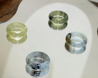 May include: Four translucent rings in shades of green, gray, and white are displayed on a reflective surface. The rings have a smooth, polished appearance, and their colors vary slightly. The rings are arranged in a square formation, with their reflections visible below.