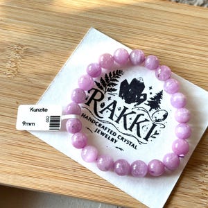 May include: A Kunzite bracelet with 9mm beads. The light purple beads are strung together, resting on a white card with the text "RAKKI HANDCRAFTED CRYSTAL JEWELRY". A price tag is attached to the card.