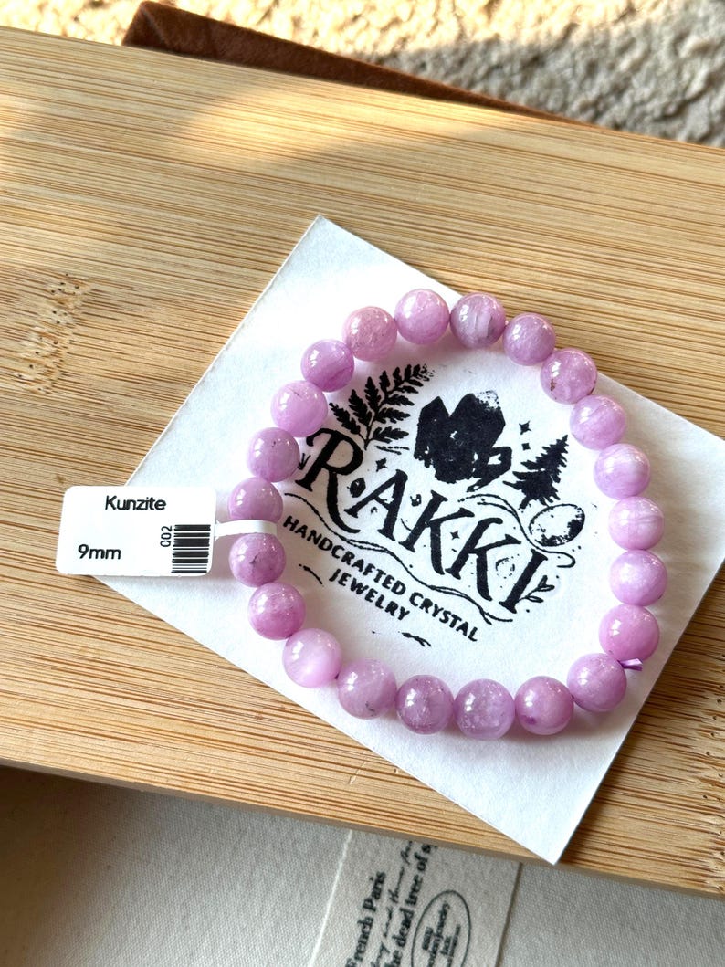 May include: A Kunzite bracelet with 9mm beads. The light purple beads are strung together, resting on a white card with the text "RAKKI HANDCRAFTED CRYSTAL JEWELRY". A price tag is attached to the card.