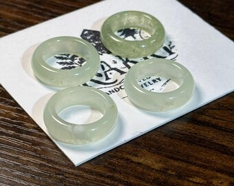 May include: Four jade rings in varying shades of green are displayed on a white card with black text. The rings are smooth and circular, with a translucent quality. The background is a dark wood surface.