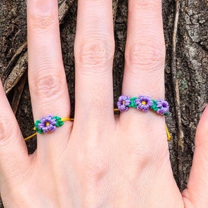 May include: A hand model wearing two purple and green flower rings on two fingers. The rings are made of string and are tied together with a yellow string.