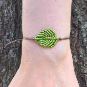 Pode incluir: Um bracelete verde em forma de folha feito de corda atada. O bracelete é usado no pulso.