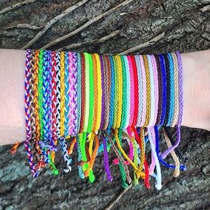 May include: A collection of colorful braided bracelets in various colors, including red, blue, green, yellow, orange, purple, pink, white, and black. The bracelets are stacked on a person's wrist.