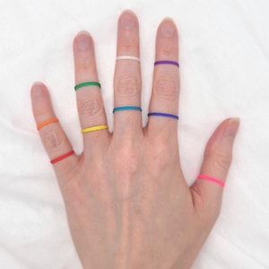 Peut inclure: Une main portant sept élastiques colorés sur différents doigts. Les élastiques sont aux couleurs de l'arc-en-ciel, en commençant par le rouge sur l'auriculaire et en terminant par le blanc sur l'index.