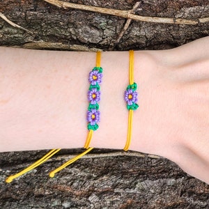May include: Two yellow friendship bracelets with purple and green flower accents. The bracelets are tied around a person's wrist.