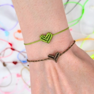 May include: Two friendship bracelets with heart-shaped knots. The top bracelet is green and the bottom bracelet is brown.