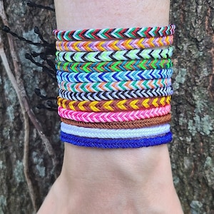May include: A person's wrist with a stack of colorful friendship bracelets. The bracelets are made of woven string and feature a chevron pattern.