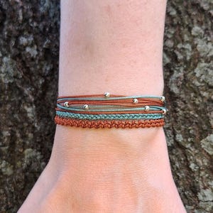 Könnte beinhalten: Ein mehrlagiges Armband mit einem geflochtenen Band und silbernen Perlen. Das Armband besteht aus einer Kombination aus braunen und blauen Bändern.