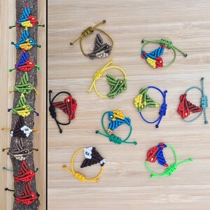 Pode incluir: Uma cole&ccedil;&atilde;o de pulseiras de amizade coloridas em forma de p&aacute;ssaro feitas com cord&atilde;o de n&oacute;. As pulseiras s&atilde;o exibidas em uma superf&iacute;cie de madeira.