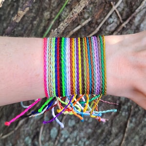 May include: A stack of colorful braided bracelets in various shades of pink, green, blue, yellow, orange, red, purple, and brown. The bracelets are tied together and worn on a person's wrist.