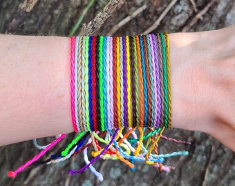 May include: A stack of colorful braided bracelets in various shades of pink, green, blue, yellow, orange, red, purple, and brown. The bracelets are tied together and worn on a person's wrist.