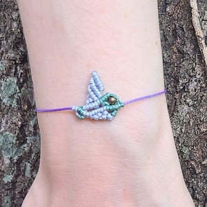 Könnte beinhalten: Ein lilafarbenes Armband aus Schnur mit einem grünen und lilafarbenen Kolibri-Anhänger. Der Anhänger hat eine kleine goldene Perle als Auge.