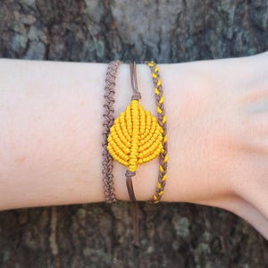 Puede incluir: Tres pulseras trenzadas marrones y amarillas con un dije de hoja amarilla. Las pulseras se llevan en la muñeca de una persona.