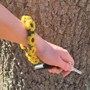 Puede incluir: Una pulsera de ganchillo hecha a mano con diseños de girasoles amarillos y hojas verdes. La pulsera se lleva en la muñeca, y la persona sostiene llaves y un llavero de coche. La pulsera está hecha de hilo suave.