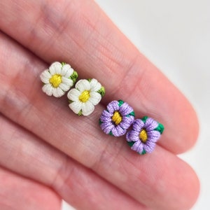 May include: A set of four handmade flower earrings. Two are white with yellow centers and green leaves, and two are purple with yellow centers and green leaves.