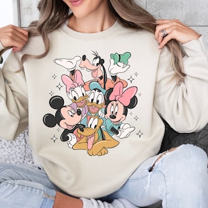 May include: Beige sweatshirt featuring a colorful graphic of Disney characters including Mickey Mouse, Minnie Mouse, Donald Duck, Daisy Duck, Goofy, and Pluto. The cartoon characters are surrounded by starbursts. The sweatshirt has a crew neck and long sleeves.