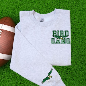 May include: A light grey sweatshirt featuring the text "BIRD GANG" in green and white, with a green and white eagle graphic. An American football is on the left. The sleeve has a green and white eagle graphic.