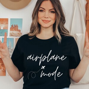Puede incluir: Camiseta negra con las palabras "airplane mode" en blanco. Un peque&ntilde;o avi&oacute;n y una l&iacute;nea de puntos est&aacute;n dibujados debajo de la palabra "airplane". La camiseta es de cuello redondo.