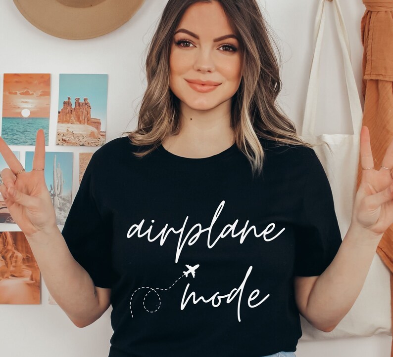 Puede incluir: Camiseta negra con las palabras "airplane mode" en blanco. Un peque&ntilde;o avi&oacute;n y una l&iacute;nea de puntos est&aacute;n dibujados debajo de la palabra "airplane". La camiseta es de cuello redondo.