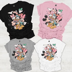 May include: Four t-shirts in black, pink, gray, and white, each featuring a colorful graphic of Disney characters including Mickey, Minnie, Donald, Daisy, Goofy, and Pluto. The design is surrounded by small star accents.