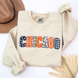May include: A beige crewneck sweatshirt featuring the word CHICAGO in large, multi-patterned block letters. Each letter displays a unique design, including navy blue with white stars, orange plaid, navy lightning bolts, orange polka dots, and navy gingham.