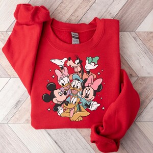 May include: A red sweatshirt featuring a colorful graphic of Disney characters including Mickey Mouse, Minnie Mouse, Donald Duck, Daisy Duck, Goofy, and Pluto. The sweatshirt is folded on a light wood-grain surface.