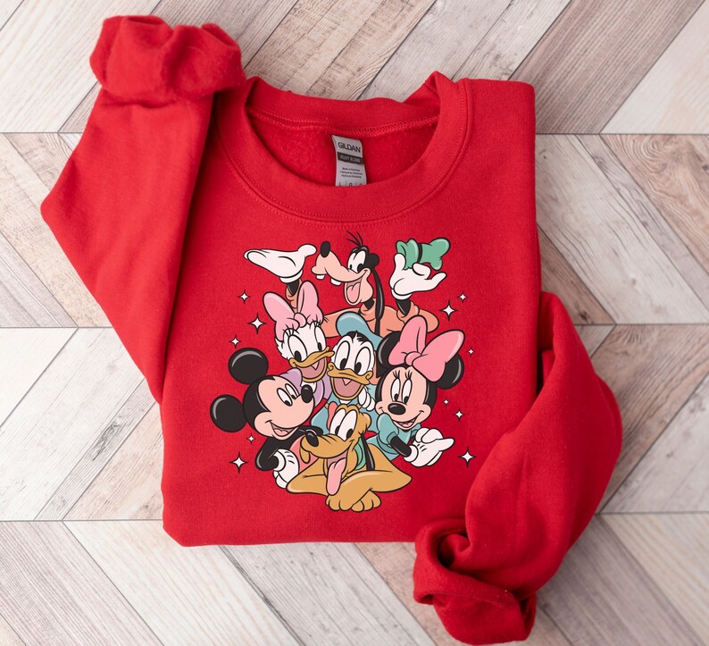 May include: A red sweatshirt featuring a colorful graphic of Disney characters including Mickey Mouse, Minnie Mouse, Donald Duck, Daisy Duck, Goofy, and Pluto. The sweatshirt is folded on a light wood-grain surface.
