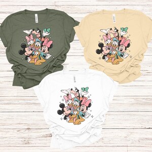 May include: Three t-shirts in olive green, cream, and white, each featuring a colorful graphic of Disney characters including Mickey Mouse, Minnie Mouse, Donald Duck, Daisy Duck, Goofy, and Pluto. The design is surrounded by small starbursts.