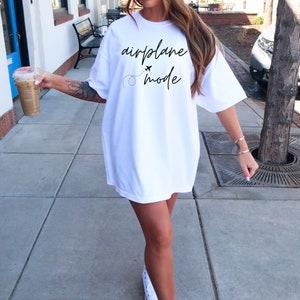 Puede incluir: Camiseta blanca de gran tama&ntilde;o con la frase "airplane mode" en escritura negra, acompa&ntilde;ada de un peque&ntilde;o gr&aacute;fico de avi&oacute;n y una trayectoria de vuelo punteada. La camiseta se lleva de forma informal.