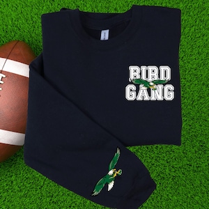 May include: Navy blue long-sleeve shirt with the text "BIRD GANG" in white block letters and a green eagle graphic. An additional eagle graphic is on the sleeve. A brown and white football is in the background.