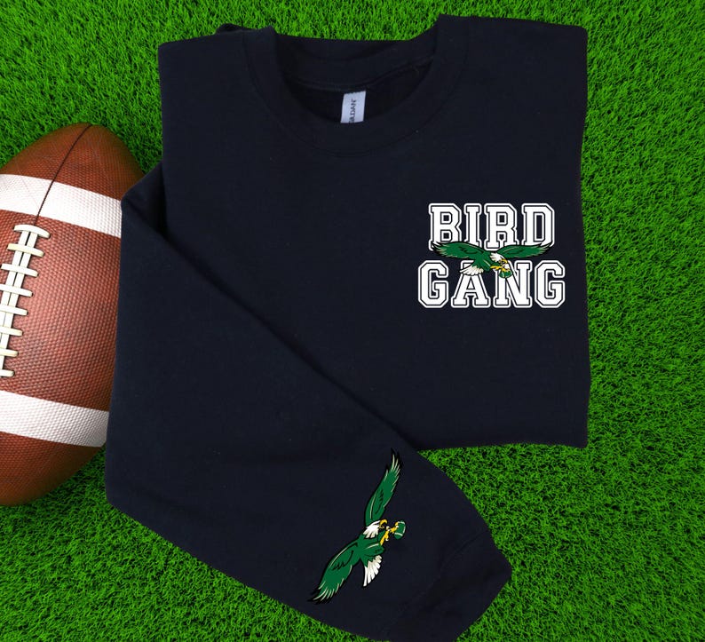 May include: Navy blue long-sleeve shirt with the text "BIRD GANG" in white block letters and a green eagle graphic. An additional eagle graphic is on the sleeve. A brown and white football is in the background.