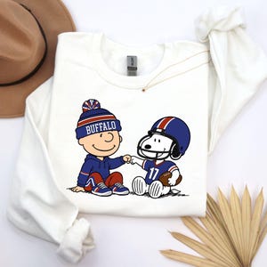 May include: A white long-sleeve sweatshirt featuring a graphic of a cartoon character wearing a blue beanie with the text BUFFALO and a cartoon dog wearing a blue football helmet with the number 17, both performing a fist bump gesture.