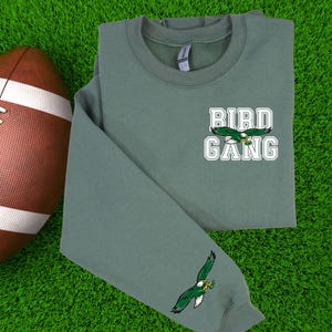 May include: A sage green sweatshirt featuring the text "BIRD GANG" and an eagle graphic. The sweatshirt has a crew neck and long sleeves, with an eagle graphic on the sleeve. A brown football is in the background.