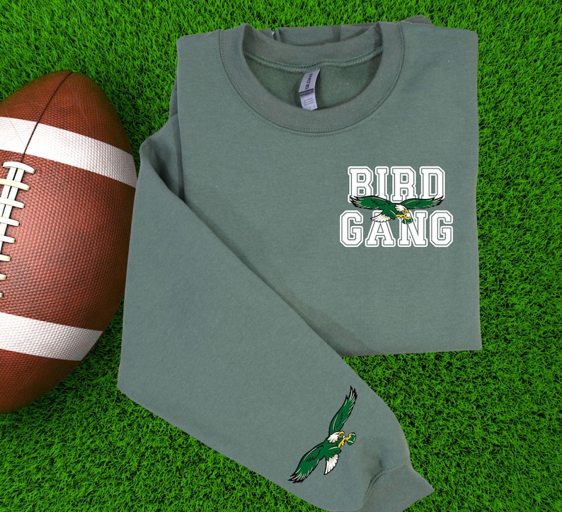 May include: A sage green sweatshirt featuring the text "BIRD GANG" and an eagle graphic. The sweatshirt has a crew neck and long sleeves, with an eagle graphic on the sleeve. A brown football is in the background.