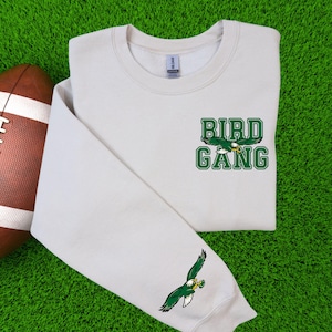 May include: A cream-coloured sweatshirt featuring the words "BIRD GANG" in green block letters and a green eagle graphic. An American football is on the left. The sleeve has a green eagle graphic.
