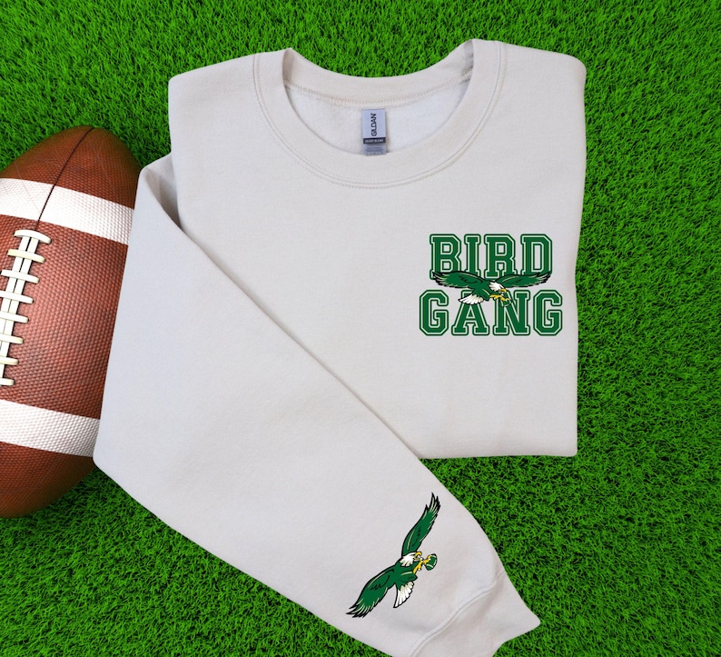 May include: A cream-coloured sweatshirt featuring the words "BIRD GANG" in green block letters and a green eagle graphic. An American football is on the left. The sleeve has a green eagle graphic.