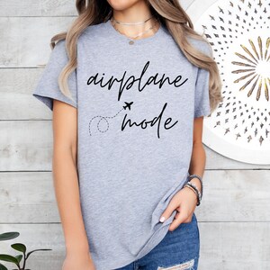 Puede incluir: Una camiseta gris claro con las palabras "airplane mode" en escritura negra. Tambi&eacute;n presenta un peque&ntilde;o gr&aacute;fico de avi&oacute;n y una l&iacute;nea de puntos. La camiseta est&aacute; hecha de un material suave.