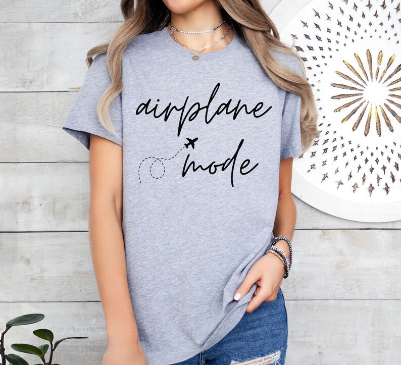 Puede incluir: Una camiseta gris claro con las palabras "airplane mode" en escritura negra. Tambi&eacute;n presenta un peque&ntilde;o gr&aacute;fico de avi&oacute;n y una l&iacute;nea de puntos. La camiseta est&aacute; hecha de un material suave.