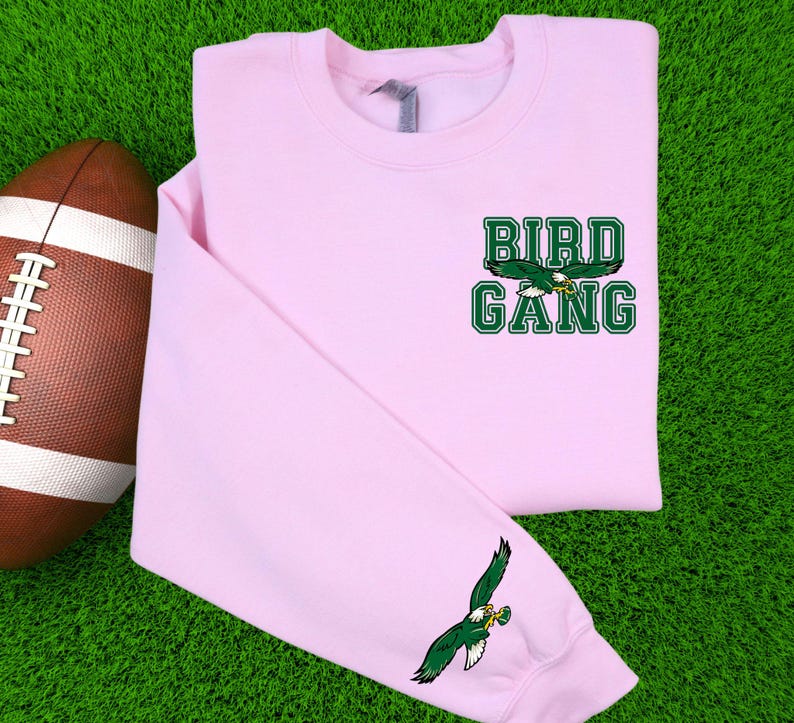 May include: A pale pink sweatshirt featuring the text "BIRD GANG" in green block letters and a green and white eagle graphic. An American football is on the left. The sleeve has a matching eagle graphic.
