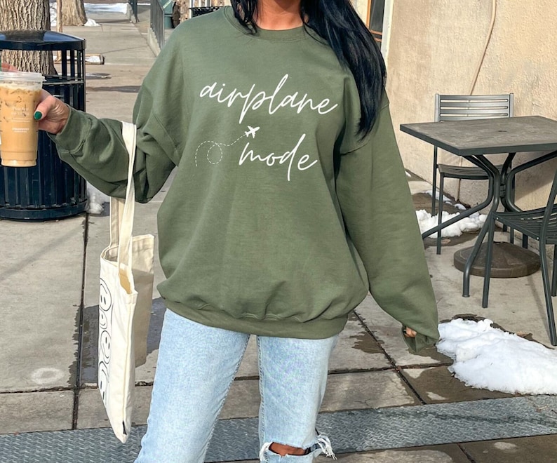 Puede incluir: Sudadera verde oliva con la frase "airplane mode" en blanco, un avi&oacute;n y una l&iacute;nea de puntos. Forma parte de una colecci&oacute;n de ropa informal.