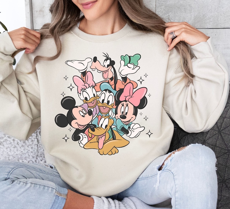 May include: Beige sweatshirt featuring a colorful graphic of Disney characters including Mickey Mouse, Minnie Mouse, Donald Duck, Daisy Duck, Goofy, and Pluto. The cartoon characters are surrounded by starbursts. The sweatshirt has a crew neck and long sleeves.