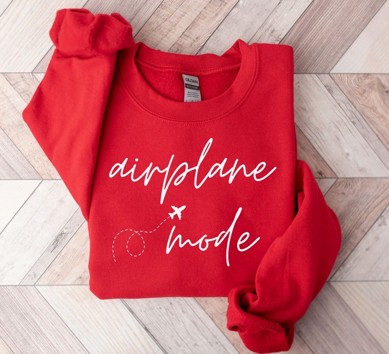 May include: A red sweatshirt with the words "airplane mode" in white script. A small airplane graphic with a dotted line trail is below the word "airplane". The sweatshirt is folded on a light wood-grain surface.