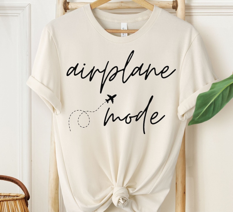 Puede incluir: Camiseta color crema con las palabras "airplane mode" en negro, con fuente manuscrita. Presenta un peque&ntilde;o gr&aacute;fico de avi&oacute;n con una l&iacute;nea de puntos. La camiseta est&aacute; anudada en la parte inferior.