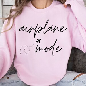 Puede incluir: Sudadera rosa claro con las palabras "airplane mode" en fuente negra. Un peque&ntilde;o avi&oacute;n y una l&iacute;nea de puntos est&aacute;n entre las palabras. La sudadera es de cuello redondo.
