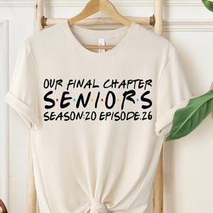May include: Cream-colored t-shirt with the text "OUR FINAL CHAPTER SENIORS SEASON:20 EPISODE:26" in black, red, yellow, and blue. The shirt is hanging on a wooden hanger.