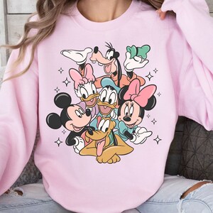 May include: A light pink sweatshirt featuring a colorful graphic of Disney characters Mickey Mouse, Minnie Mouse, Donald Duck, Daisy Duck, Goofy, and Pluto. The design includes star accents.