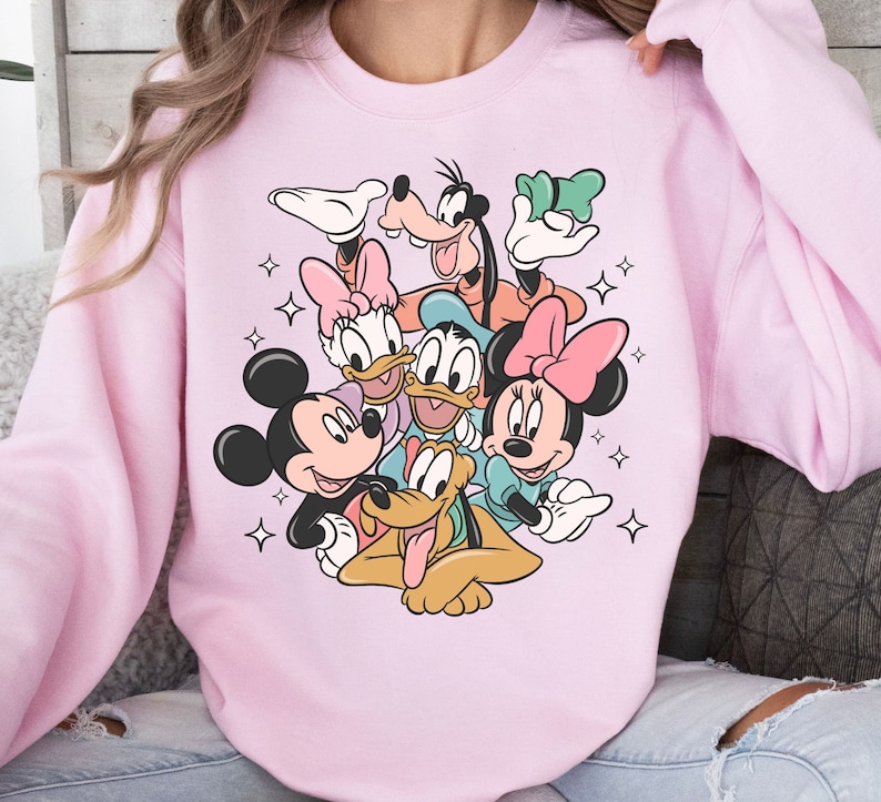 May include: A light pink sweatshirt featuring a colorful graphic of Disney characters Mickey Mouse, Minnie Mouse, Donald Duck, Daisy Duck, Goofy, and Pluto. The design includes star accents.
