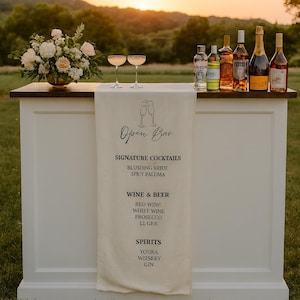 Puede incluir: Un bar al aire libre con una encimera blanca y una tapa de madera oscura. El bar cuenta con un arreglo floral, copas de champán y varias botellas de licor. Una pancarta muestra "Open Bar" y opciones de cócteles.